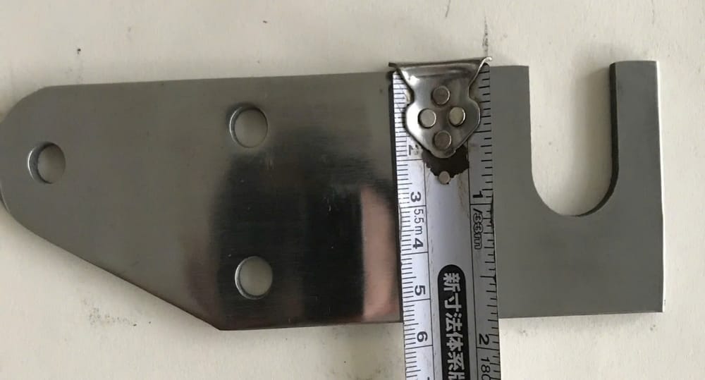 Pát thẳng inox bản mã ba lỗ bắt vít, một đầu vát và một đầu xẻ rãnh chữ U, đặt cạnh thước dây để đối chiếu kích thước