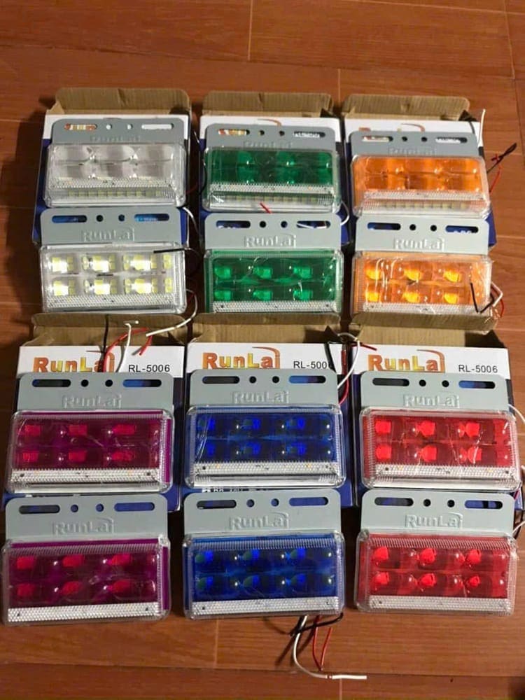 Mười hai đèn định vị mạng sườn LED xếp thành hàng trên sàn kho, đủ các màu trắng, xanh lá, vàng, đỏ và xanh dương, mỗi đèn có đế bắt vít và hai dây nguồn